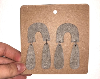 May include: A pair of gray speckled teardrop-shaped earrings with a small arch at the top. The earrings are displayed on a brown card.