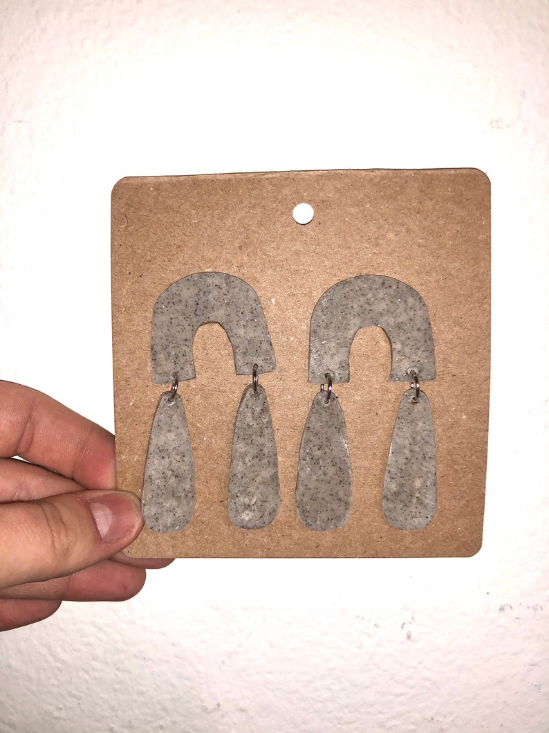 May include: A pair of grey speckled teardrop-shaped earrings with a small arch at the top. The earrings are displayed on a brown card.
