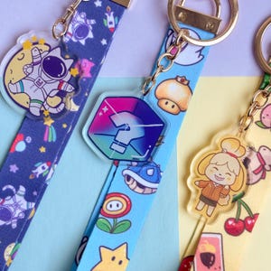 May include: Close-up of three keychains with colorful lanyards. One lanyard is navy blue with space-themed graphics and an astronaut charm. Another is light blue with video game characters and a question mark charm. The third is yellow with cherries and a cartoon character.