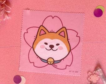 May include: A pink microfiber cleaning cloth with a cartoon Shiba Inu dog wearing a blue collar with a bell. The dog is surrounded by a pink cherry blossom flower. The text "GORI" and "ART BY GORI" are printed on the cloth.