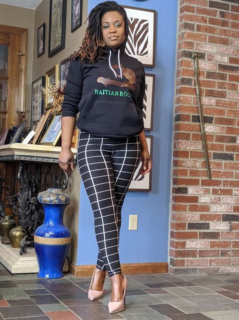 May include: A woman wearing a black hoodie with a graphic of the island of Haiti and the text "Haitian Roo" in green. She is also wearing black and white checkered leggings and pink high heels.