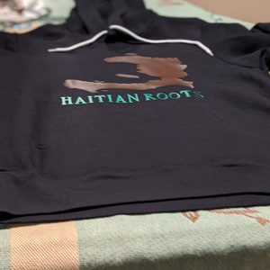May include: Black hooded sweatshirt with a brown outline of the island of Haiti and the text "HAITIAN ROOTS" in green.