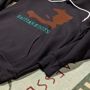 May include: Black hooded sweatshirt with a brown outline of Haiti and the text "Haitian Roots" in teal green.