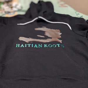 May include: Black hooded sweatshirt with a brown outline of the island of Haiti and the text "HAITIAN ROOTS" in teal green.
