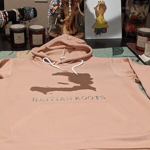 May include: A peach-colored hoodie with a white drawstring and a brown outline of Haiti on the front. The text "HAITIAN KOOTS" is printed in silver below the outline.