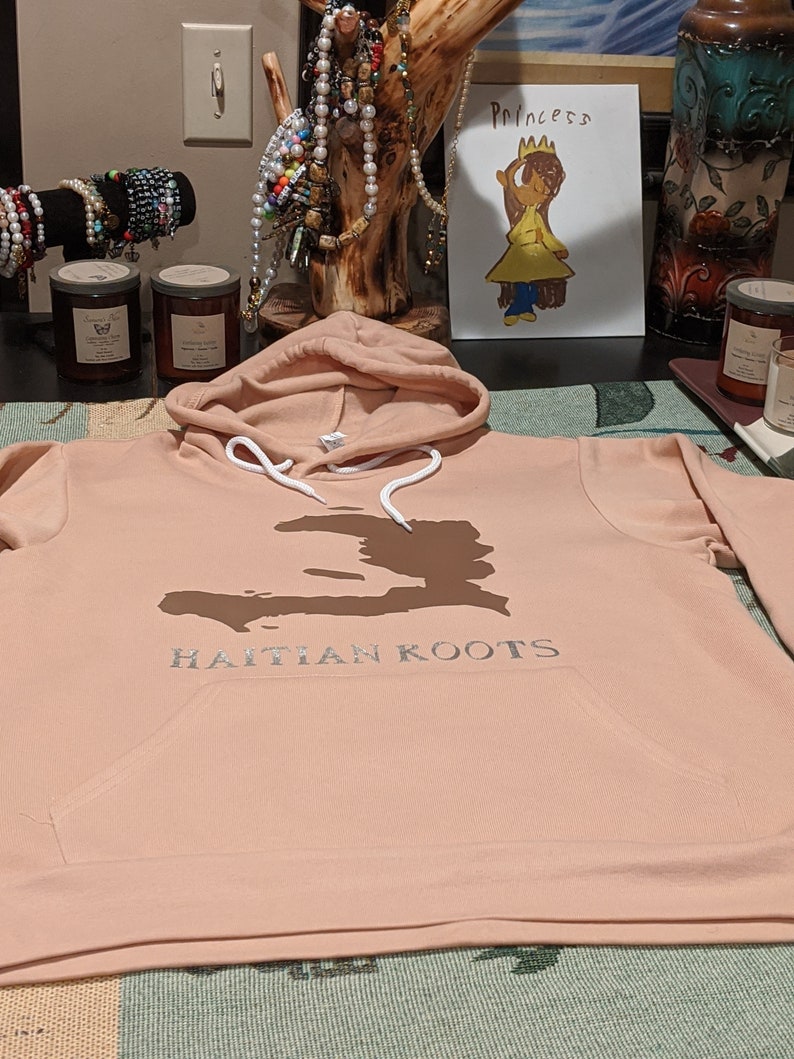May include: A peach-colored hoodie with a white drawstring and a brown outline of Haiti on the front. The text "HAITIAN KOOTS" is printed in silver below the outline.