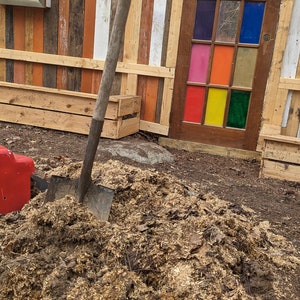 May include: A metal shovel with a wooden handle is partially buried in a pile of wood chips. The shovel is in front of a wooden wall with a colorful glass door and a mirror.