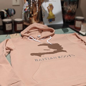 May include: A light pink hoodie with a brown outline of Haiti and the text "HAITIAN ROOTS" printed on the front.