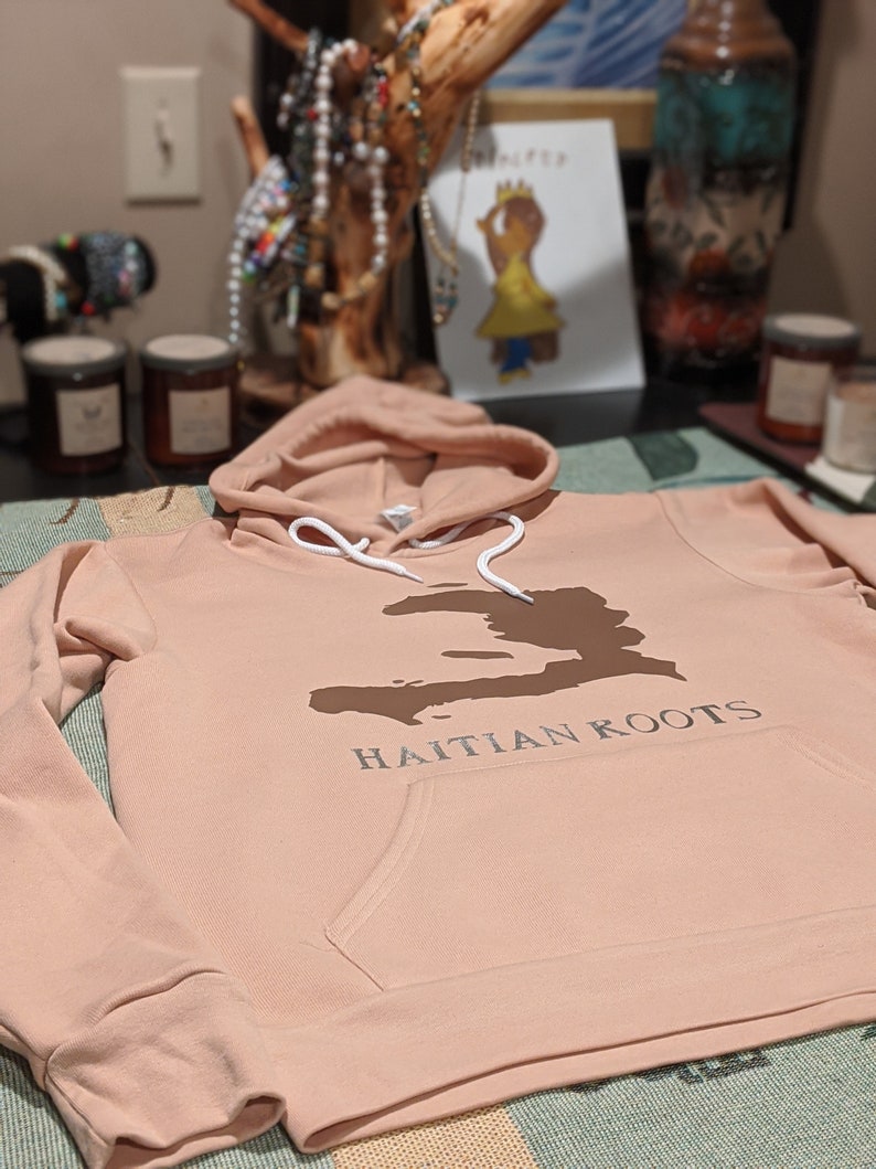 May include: A light pink hoodie with a brown outline of Haiti and the text "HAITIAN ROOTS" printed on the front.