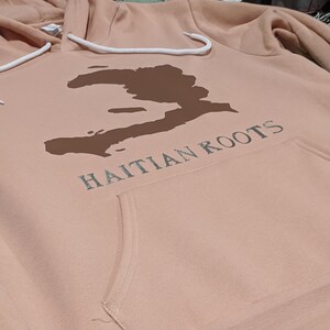 May include: A light pink hoodie with a brown outline of Haiti and the text "Haitian Roots" in white.