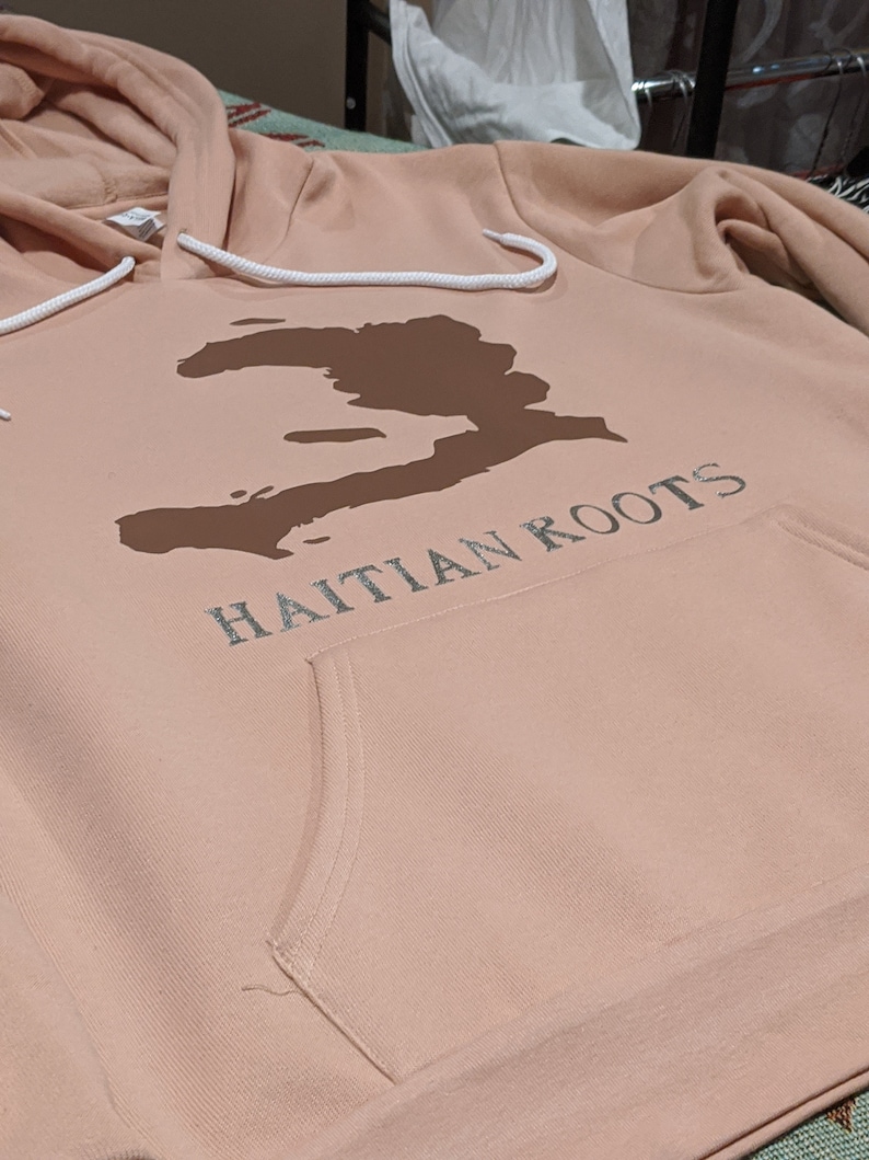 May include: A light pink hoodie with a brown outline of Haiti and the text "Haitian Roots" in white.