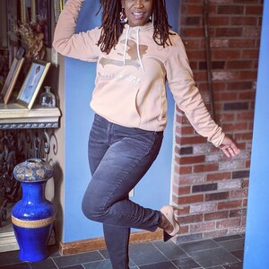 May include: A woman wearing a peach-colored hoodie with a brown outline of the island of Hispaniola and the text "Haitian Roots". She is also wearing black jeans and brown heeled shoes.