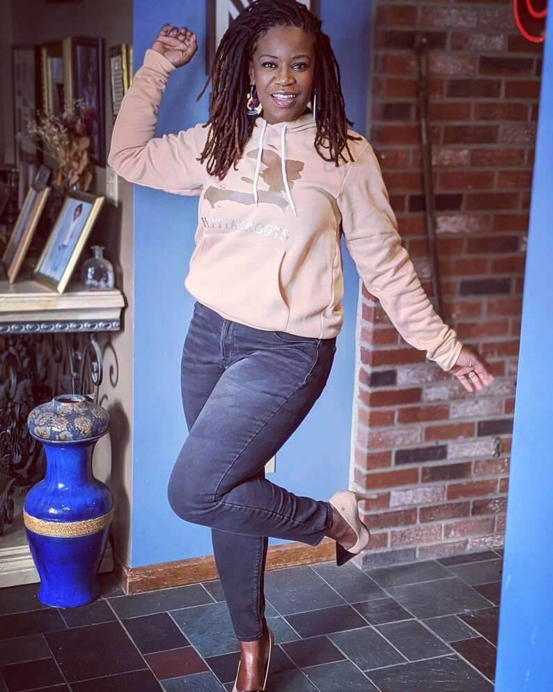 May include: A woman wearing a peach-colored hoodie with a brown outline of the island of Hispaniola and the text "Haitian Roots". She is also wearing black jeans and brown heeled shoes.