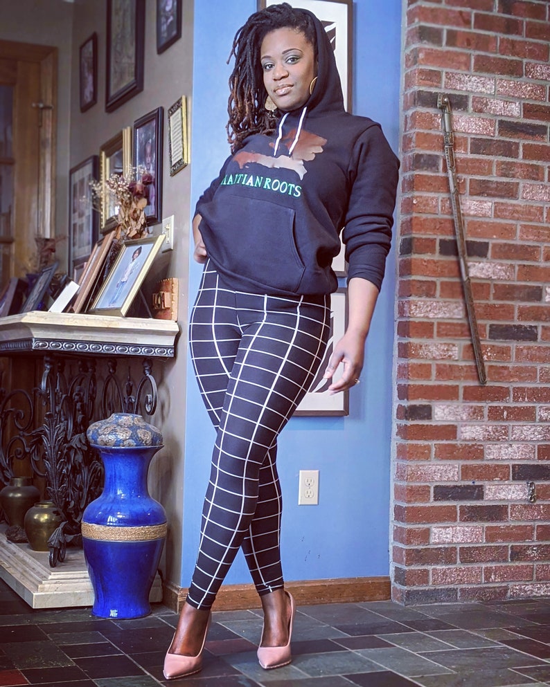 May include: A woman wearing a black hoodie with the text "Haitian Roots" and a graphic of the island of Hispaniola. She is also wearing black and white checkered leggings and pink heels.