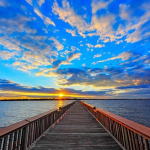 May include: A wooden pier extends out into a body of water, with a bright blue sky and fluffy white clouds overhead. The sun is setting in the distance, casting a warm glow on the water.