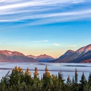 Peut inclure: Un paysage pittoresque avec un plan d'eau entouré de montagnes sous un ciel bleu et blanc strié. Le premier plan présente des arbres verts, avec des montagnes enneigées.