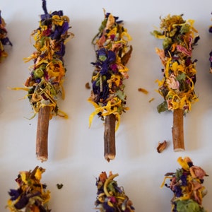 K&ouml;nnte beinhalten: Draufsicht auf mehrere Holzst&auml;be, die mit getrockneten Blumen verziert sind. Die Blumen sind eine Mischung aus Gelb, Lila und Rosa und erzeugen ein farbenfrohes und strukturiertes Aussehen. Die St&auml;be sind auf einer wei&szlig;en Oberfl&auml;che angeordnet.