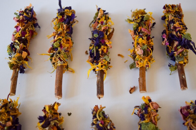 K&ouml;nnte beinhalten: Draufsicht auf mehrere Holzst&auml;be, die mit getrockneten Blumen verziert sind. Die Blumen sind eine Mischung aus Gelb, Lila und Rosa und erzeugen ein farbenfrohes und strukturiertes Aussehen. Die St&auml;be sind auf einer wei&szlig;en Oberfl&auml;che angeordnet.