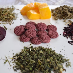 Pu&ograve; includere: Una superficie bianca con varie erbe secche e ingredienti, tra cui fette di barbabietola rossa, fiori di ibisco secchi, pezzi di mango e piccoli biscotti di t&egrave; pressati rossi.