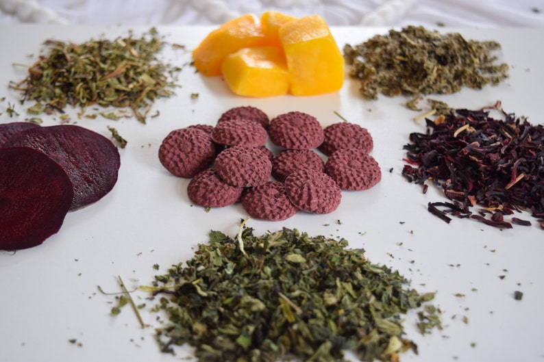 Pu&ograve; includere: Una superficie bianca con varie erbe secche e ingredienti, tra cui fette di barbabietola rossa, fiori di ibisco secchi, pezzi di mango e piccoli biscotti di t&egrave; pressati rossi.