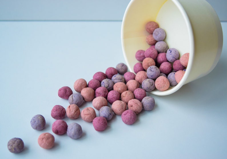 K&ouml;nnte beinhalten: Eine hellgelbe Schale, die auf der Seite liegt und zahlreiche kleine, kugelf&ouml;rmige Objekte in Rosa-, Lila- und Lavendelt&ouml;nen versch&uuml;ttet. Die runden Gegenst&auml;nde haben eine texturierte, matte Oberfl&auml;che und liegen vor der Schale verstreut.