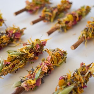 K&ouml;nnte beinhalten: Draufsicht auf mehrere R&auml;ucherb&uuml;ndel, jedes mit einem Holzsockel, verziert mit getrockneten Blumen und Kr&auml;utern. Die Anordnung umfasst eine Vielzahl von Farben, wie Gelb, Rosa und Gr&uuml;n, was eine optisch ansprechende und nat&uuml;rliche &Auml;sthetik erzeugt.