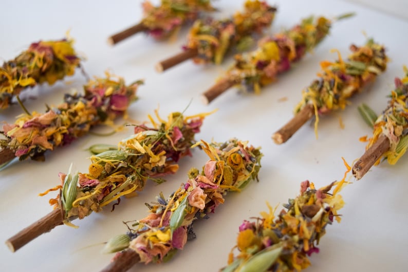 K&ouml;nnte beinhalten: Draufsicht auf mehrere R&auml;ucherb&uuml;ndel, jedes mit einem Holzsockel, verziert mit getrockneten Blumen und Kr&auml;utern. Die Anordnung umfasst eine Vielzahl von Farben, wie Gelb, Rosa und Gr&uuml;n, was eine optisch ansprechende und nat&uuml;rliche &Auml;sthetik erzeugt.