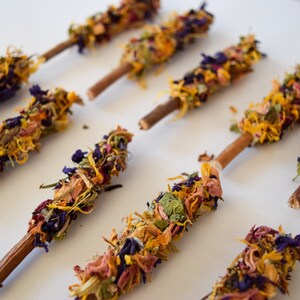 K&ouml;nnte beinhalten: Nahaufnahme von mehreren Blumenst&auml;ben. Jeder Stab hat einen Holzstab als Basis, der mit einer Vielzahl von getrockneten Blumen in Gelb-, Orange-, Lila- und Rosat&ouml;nen bedeckt ist. Die St&auml;be sind auf einer wei&szlig;en Oberfl&auml;che angeordnet.