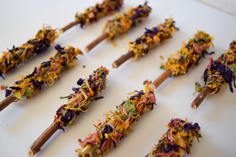 K&ouml;nnte beinhalten: Nahaufnahme von mehreren Blumenst&auml;ben. Jeder Stab hat einen Holzstab als Basis, der mit einer Vielzahl von getrockneten Blumen in Gelb-, Orange-, Lila- und Rosat&ouml;nen bedeckt ist. Die St&auml;be sind auf einer wei&szlig;en Oberfl&auml;che angeordnet.
