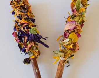 Può includere: Due bastoncini di fiori secchi per fumigazione. I bastoncini sono avvolti con una varietà di fiori secchi colorati, tra cui petali viola, arancioni, gialli e rosa. I bastoncini hanno un manico in legno naturale.