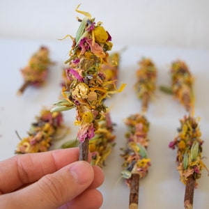 K&ouml;nnte beinhalten: Eine Sammlung von Trockenblumen-B&uuml;ndeln, die jeweils an einem kleinen Holzstab befestigt sind. Die B&uuml;ndel weisen eine Mischung aus gelben, rosa und gr&uuml;nen Trockenblumen und Kr&auml;utern auf. Der Hintergrund ist eine schlichte wei&szlig;e Oberfl&auml;che.
