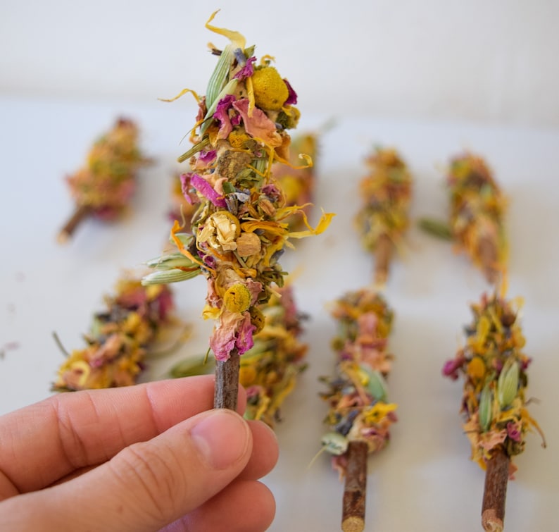 K&ouml;nnte beinhalten: Eine Sammlung von Trockenblumen-B&uuml;ndeln, die jeweils an einem kleinen Holzstab befestigt sind. Die B&uuml;ndel weisen eine Mischung aus gelben, rosa und gr&uuml;nen Trockenblumen und Kr&auml;utern auf. Der Hintergrund ist eine schlichte wei&szlig;e Oberfl&auml;che.