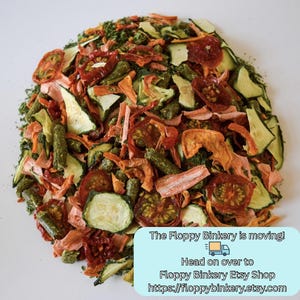 May include: A mixture of dried vegetable pieces including sliced green courgette, red tomato slices, orange carrot strips, and small green cylindrical pellets. A light blue rectangular sign with text about a shop move is in the bottom right corner.