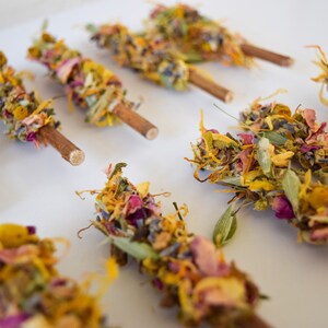 K&ouml;nnte beinhalten: Nahaufnahme von mehreren Trockenblumen-B&uuml;ndeln auf Holzst&auml;ben. Die B&uuml;ndel bestehen aus bunten getrockneten Bl&uuml;tenbl&auml;ttern, Bl&auml;ttern und Knospen in Gelb-, Rosa-, Lila- und Gr&uuml;nt&ouml;nen. Die St&auml;be sind naturbraun.