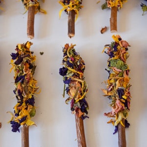K&ouml;nnte beinhalten: Nahaufnahme von mehreren Holzst&auml;ben, die mit getrockneten Blumen in Gelb-, Lila-, Rosa- und Gr&uuml;nt&ouml;nen verziert sind. Die floralen Arrangements sind oben auf jedem Stab angeordnet und erzeugen eine dekorative und nat&uuml;rliche &Auml;sthetik.