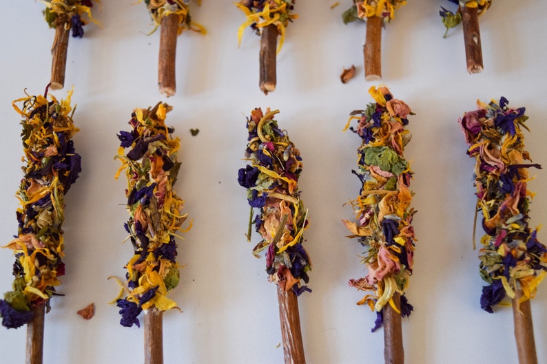 K&ouml;nnte beinhalten: Nahaufnahme von mehreren Holzst&auml;ben, die mit getrockneten Blumen in Gelb-, Lila-, Rosa- und Gr&uuml;nt&ouml;nen verziert sind. Die floralen Arrangements sind oben auf jedem Stab angeordnet und erzeugen eine dekorative und nat&uuml;rliche &Auml;sthetik.
