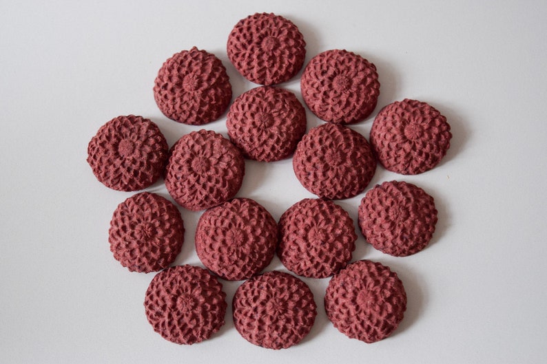 Pu&ograve; includere: Una collezione di 15 bottoni decorativi rotondi di colore bordeaux con un design floreale in rilievo. I bottoni sono disposti in cerchio su uno sfondo bianco.
