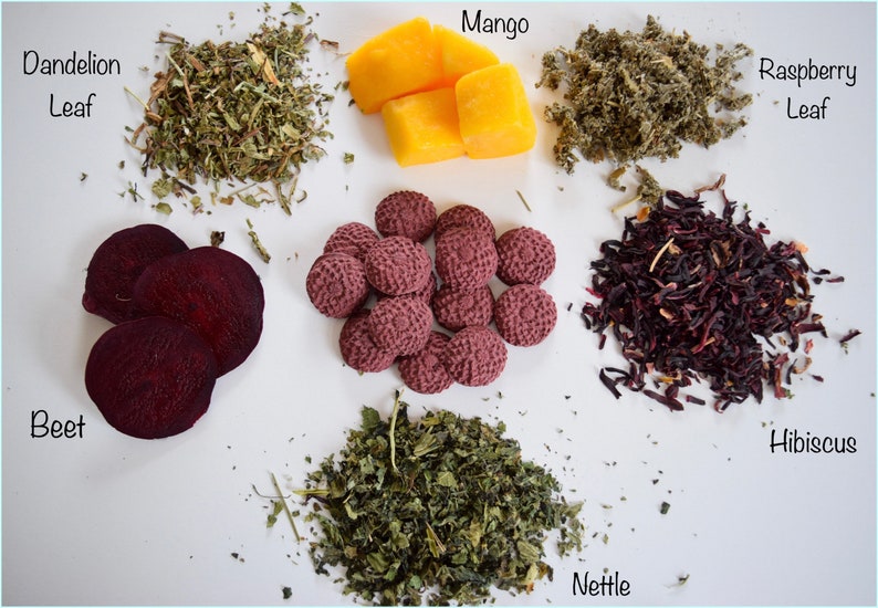 Pu&ograve; includere: Una variet&agrave; di erbe e frutti secchi, tra cui foglie di tarassaco, foglie di lampone, ortica, ibisco, barbabietola e mango. Gli ingredienti sono disposti su una superficie bianca.