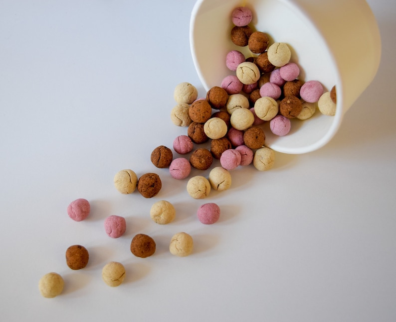K&ouml;nnte beinhalten: Eine wei&szlig;e Papierschale, die auf der Seite liegt und kleine, runde, texturierte Kugeln in Beige-, Braun- und Hellrosat&ouml;nen auf eine glatte Fl&auml;che sch&uuml;ttet. Jede Kugel weist einen deutlichen Riss auf, was auf eine gebackene oder getrocknete Textur hindeutet.