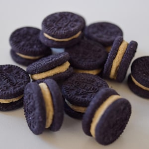 May include: A pile of dark purple sandwich cookies with a light tan creme filling. The cookies are round with a textured surface and a logo imprint. The cookies are stacked and scattered on a white surface.