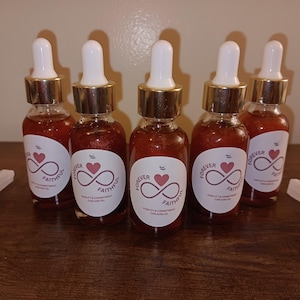 Könnte beinhalten: Fünf bernsteinfarbene Glasflaschen mit goldenen Deckeln und Etiketten mit der Aufschrift "Forever Faithful" mit einem roten Herzen und einem Unendlichkeitszeichen. Die Flaschen stehen auf einer Holzoberfläche.