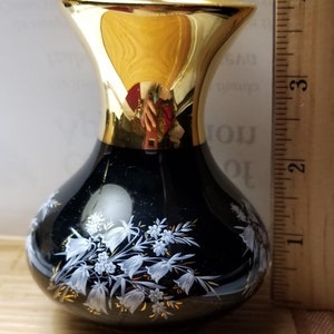 May include: A small, black glass vase with a gold rim and floral design. The vase is approximately 3 inches tall. White floral patterns with gold accents adorn the vase's surface, creating a decorative piece for home decor.