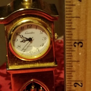 May include: A gold-colored miniature grandfather clock with a white face and black hands. The clock is about 4 inches tall and has a red base. The clock face says "Quartz" and has black Roman numeral markings.