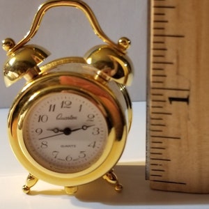 May include: A small, gold-colored Quartz alarm clock with a white face and black numerals. The clock has a curved handle and two bells on top. A wooden ruler is next to the clock.