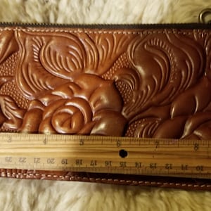 May include: A brown leather wallet with a floral embossed design. The wallet is closed and has a zipper closure. A ruler is placed next to the wallet to show its size.