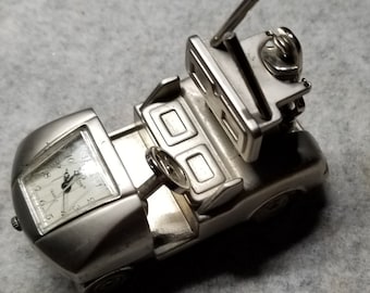 May include: A silver-colored golf cart clock with a black face and white numbers. The clock is in the shape of a golf cart with a steering wheel and a golf club sticking up.