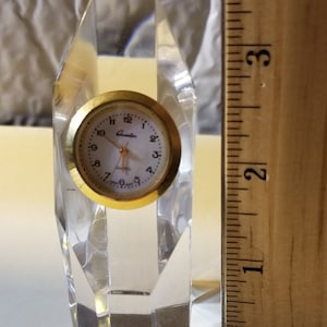May include: A small, clear crystal clock with a gold-tone face and black hands. The clock is about 4 inches tall and has a quartz movement. The clock is made in China.