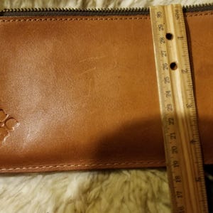 May include: A brown leather wallet with a zipper closure and a small flower embossed on the front. The wallet is 7 inches long.