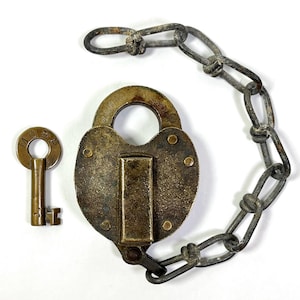 May include: A heart-shaped brass padlock with a key and chain. The padlock has a brass keyhole and a brass hasp. The key is made of brass and has a round head with the letters "M.C.E.R." engraved on it. The chain is made of iron and is attached to the padlock with a loop.