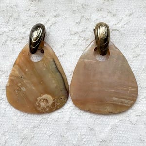 May include: A pair of earrings featuring large, triangular, translucent tan shell pendants suspended from metallic, oval-shaped gold-tone clips. The shell pieces exhibit natural variations in color, texture, and pattern, with a smooth, polished finish.